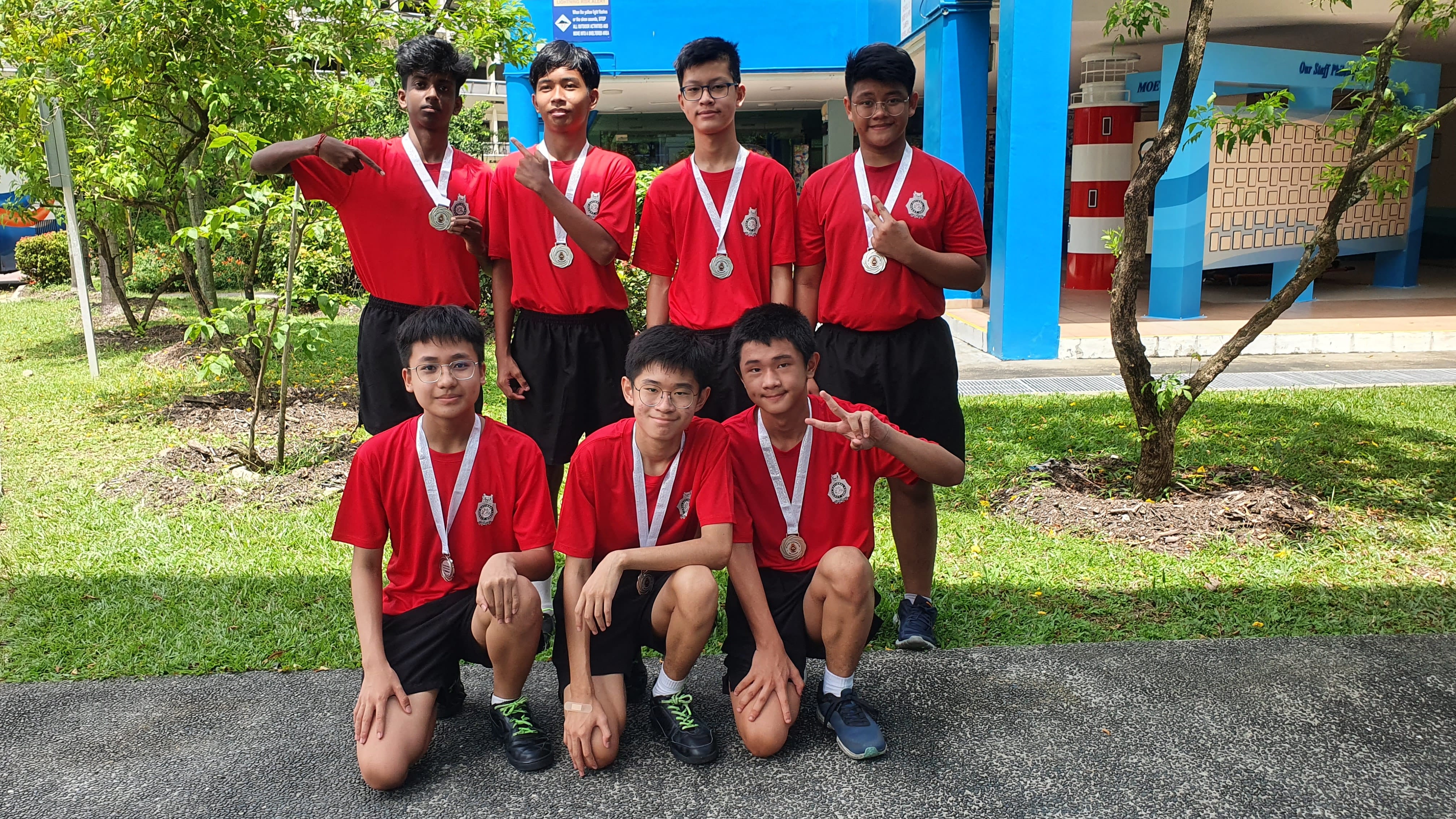 Seven teenage boys in red shirts and black shorts wear medals, posing in two rows outdoors near a blue building.
